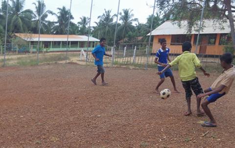 Four boys are kicking the soccer ball on the dirt ground.