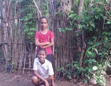 A boy kneeling on the ground and behind him is a girl standing in front of a stick fence.