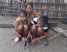 Three children hold a chicken, which is eating rice on the ground. 